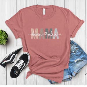 May include: A dusty rose t-shirt with the word "MAMA" in block letters and the names "Kevin" and "Alice" in a script font. The shirt is paired with denim shorts and black and white sneakers.