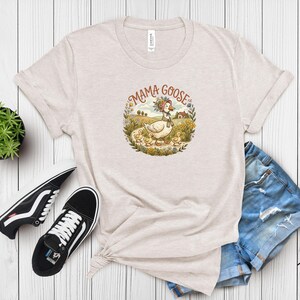 May include: Beige t-shirt featuring a circular graphic with the words "Mama Goose." The graphic depicts a goose wearing a floral crown and scarf, walking with her goslings through a field of flowers. The shirt is paired with denim shorts and black sneakers.