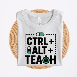 May include: A white t-shirt featuring a black graphic print that reads CTRL + ALT + TEACH. The design includes a green toggle switch icon at the top, a green toggle switch icon on the letter A, and a green power button icon on the letter A.