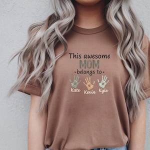 May include: A brown t-shirt with the text "This awesome MOM belongs to" above three handprints. The handprints are in shades of green and orange, with the names Kate, Kevin, and Kyle below.