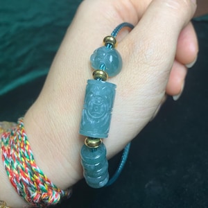 Può includere: Un braccialetto di giada con un cordino verde acqua e dettagli color oro. Il braccialetto presenta perline di giada scolpite a forma di fiore, un cilindro con un design dettagliato e anelli impilati. La giada è traslucida.