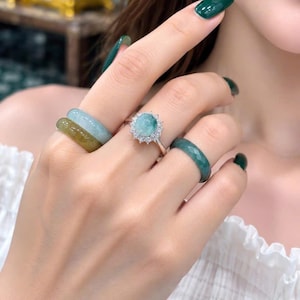 Op de afbeelding: Een set van vier ringen gedragen aan de vingers. Twee ringen zijn gladde, massieve banden in tinten lichtgroen en olijfgroen. Eén ring heeft een centrale ronde groene steen omringd door een halo van kleine heldere steentjes. Eén ring is gefacetteerd.