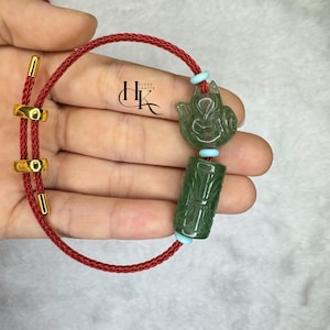 May include: A jade bracelet with a red cord and gold-colored accents. The bracelet features two carved jade pendants: a fox and a cylindrical piece with carved details. The bracelet is held in a hand.