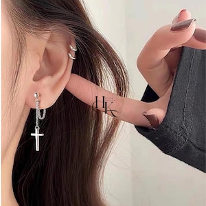 May include: A silver earring with a dangling cross design. The earring features a small ball stud, a chain, and a cross pendant. The image also shows a double hoop earring and the text "HK STONE CRAFTS".