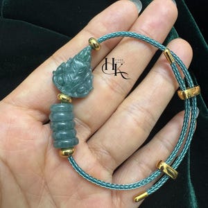 May include: A jade bracelet featuring a carved green pendant and stacked green beads. The bracelet has a teal cord with gold-colored accents. The bracelet is held in a hand.