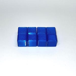Pode incluir: Um conjunto de oito pequenos cubos de cor azul vibrante dispostos numa grelha retangular de dois por quatro. Cada cubo individual apresenta uma superfície lisa e uniforme com texturas em camadas visíveis, formando um bloco coeso.