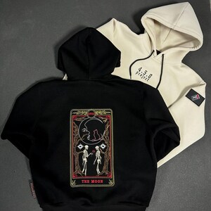 Pode incluir: Duas camisolas com capuz, uma preta e outra creme, são exibidas. A camisola preta apresenta um desenho de carta de tarot nas costas com a inscrição "THE MOON". A camisola creme tem o texto "4.5.0 project" na frente.