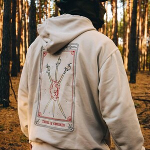 Könnte beinhalten: Beiger Kapuzenpullover mit Tarotkarten-Design auf dem Rücken. Die Karte zeigt drei Schwerter, die ein Herz durchbohren, mit den Worten "THREE OF SWORDS" darunter. Der Kapuzenpullover wird in einem Wald getragen.