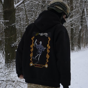 May include: Black hooded sweatshirt with a graphic on the back featuring a skeleton figure with a scythe, framed in gold. The person is wearing camouflage pants and a helmet, standing in a snowy forest.