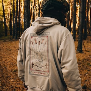 Könnte beinhalten: Beige Kapuzenpullover mit einem Tarotkarten-Design auf dem Rücken. Die Grafik zeigt einen Schädel, Schwerter und den Text "NINE OF SWORDS" in einem rot-beigen Farbschema. Die Person trägt einen Tarnhelm.
