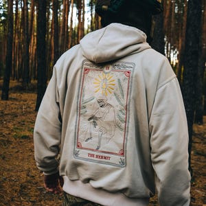 Può includere: Felpa con cappuccio beige con un grande disegno ricamato di carta dei tarocchi sul retro. La carta presenta un'illustrazione di una figura in abito con un bastone, il testo "THE HERMIT" e il numero IX. La felpa ha un cappuccio e un orlo a coste.