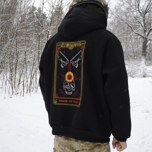 May include: Black hoodie with a tarot card design on the back. The card features a skull, sunflower, and crossed bones, with the text "THREE OF CUPS". The person is wearing camouflage pants and a helmet.