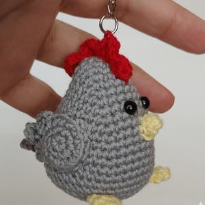 May include: A handmade crochet chicken keyring. The chicken is mainly grey with a red comb, black eyes, and yellow beak and feet. The keyring is held by a silver metal ring.