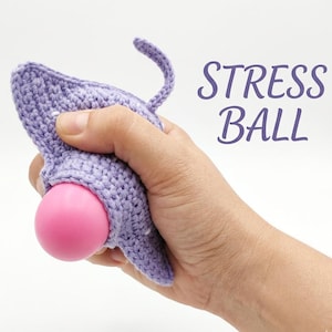 May include: A lavender crocheted stress ball with a pink sphere peeking out. The stress ball is held in a hand. The words "STRESS BALL" are written in a cursive font in the upper right corner.