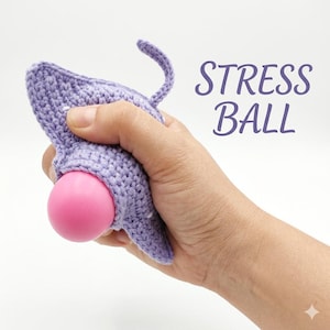 Pode incluir: Uma bola de stress de crochê lavanda em forma de gato, com uma bola rosa visível. O texto "STRESS BALL" está em roxo no lado direito. A cauda do gato está enrolada.