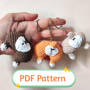 Op de afbeelding: Drie gehaakte luiaard sleutelhangers in bruin, oranje en beige, elk met een wit gezicht en poten, en een zwarte gestikte mond en oogdetail. De sleutelhangers zijn bevestigd aan zilveren metalen ringen. De afbeelding bevat de tekst "PDF Pattern".