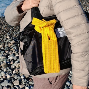 May include: A vibrant yellow knitted scarf is draped over a black bag. The scarf features a ribbed texture and is knotted at the top. The bag has a tag that reads "Hengla". The background shows a person wearing a beige jacket.