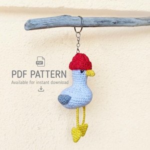 May include: A crochet seagull keyring with a light blue body, a red hat, a yellow beak, and yellow feet. The seagull hangs from a metal ring. Text on the left reads PDF PATTERN Available for instant download with a PDF icon and arrow.