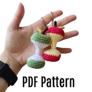 May include: Two handmade crochet keyrings shaped like apples, one green and white, the other red and yellow. Each keyring has a silver ring and a brown stem. The text "PDF Pattern" is displayed in black.