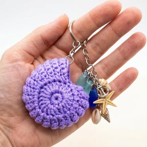 May include: A handmade, crochet keychain featuring a lavender seashell design. The keychain includes a silver ring and chain, with charms of sea glass, seashells, a starfish, and an anchor. The seashell is approximately 3 inches wide.
