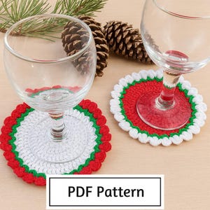Pode incluir: Duas taças de vinho repousam sobre bases de crochê feitas à mão. As bases apresentam um centro branco, ladeado por verde e vermelho, com uma borda ondulada. A imagem inclui pinhas e o texto "PDF Pattern".