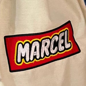 Könnte beinhalten: Cremefarbener Kordelzugbeutel mit blauen Seilriemen. Der Beutel hat einen rechteckigen Aufnäher mit dem Namen "MARCEL" in weißen Buchstaben auf rotem Grund mit gelbem Rand. Der Beutel ist aus robustem Stoff.