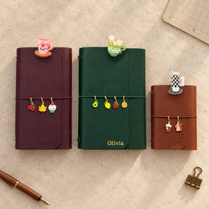 May include: Three leather journals in burgundy, dark green, and brown. The burgundy journal features the name Emily, the green one says Olivia, and the brown one says FY. Each journal is adorned with colorful charms and a decorative clip.