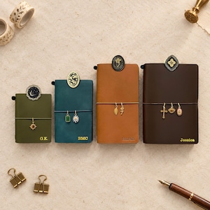 May include: Four leather journals in olive, teal, tan, and dark brown, each featuring a personalized name or initials, a decorative metal clip at the top, and dangling charms attached to an elastic band. The journals vary in size.