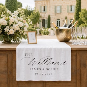 Peut inclure: Un chemin de table en lin blanc avec le texte imprimé en noir THE Williams JAMES & SOPHIA 08.12.2026. Le chemin est posé sur une table en bois avec un bol en métal martelé doré contenant des bouteilles de champagne et des flûtes en verre.