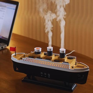 Op de afbeelding: Een scheepsvormige essentiële olie diffuser in zwart, wit en bruin, met drie schoorstenen die stoom uitstoten. Het model heeft een rode vlag en een houten dek. De diffuser staat op een houten oppervlak.
