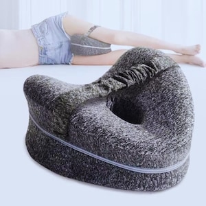 May include: A grey and black contoured leg pillow with a zip closure. The pillow has a curved design with a central opening. The image shows the pillow in use, positioned between the legs.