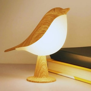 Könnte beinhalten: Eine vogelförmige Tischleuchte mit einer hellen Holzmaserung an Kopf, Rücken und Sockel sowie einem leuchtend weißen, beleuchteten Brustbereich. Die Lampe steht auf einem runden Fuß und strahlt ein sanftes Licht aus ihrem Körper aus.