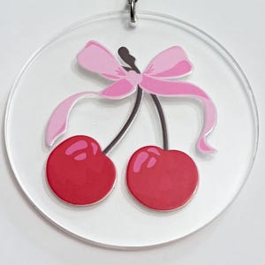 May include: A clear acrylic keychain featuring a pair of red cherries with a pink bow. The cherries have a glossy finish and are connected by a brown stem. The keychain is attached to a silver metal ring.
