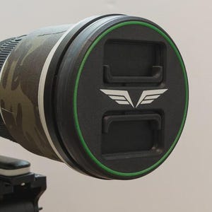 May include: A camera lens with a camouflage sleeve and a black lens cap. The lens cap has a green trim and a white winged logo. The lens is mounted on a tripod.