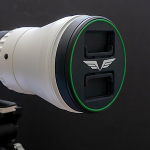 Peut inclure: Un objectif de téléphoto blanc et noir avec un bouchon d'objectif noir avec un bord vert et un logo blanc. L'objectif est monté sur un trépied noir, sur fond sombre.