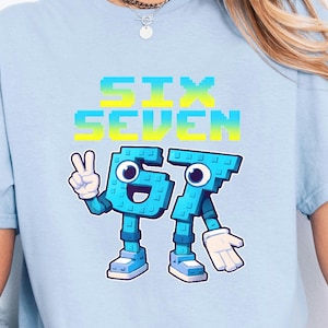 May include: Light blue t-shirt with the text "SIX SEVEN" in a gradient of blue and yellow. The graphic features the numbers 6 and 7 as cartoon characters. The number 6 is giving a peace sign. The shirt is made of a soft material.