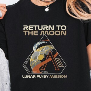 May include: Black t-shirt with a graphic design featuring the text "RETURN TO THE MOON" above a triangular emblem. The emblem includes a stylized moon and the words "LUNAR FLYBY MISSION".