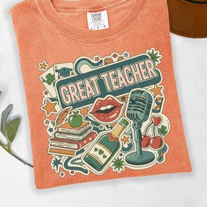 May include: Orange Comfort Colors t-shirt with a colorful graphic that says "GREAT TEACHER." The graphic includes a microphone, lips, books, a champagne bottle, and other teacher-related icons. A brown hat is partially visible.