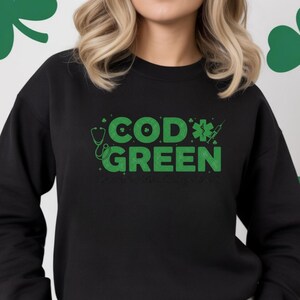May include: Black sweatshirt with the words "COD GREEN" in green, along with a stethoscope and syringe graphic. The sweatshirt is worn by a person with blonde hair and sunglasses. The background has green shamrock decorations.