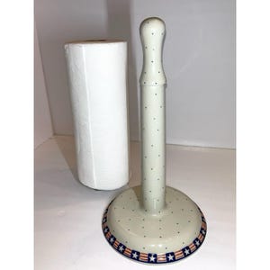 May include: A ceramic paper towel holder with a cream-coloured base and vertical post, decorated with small dark blue dots. The circular base features a decorative border pattern of alternating red and white stripes with blue stars along the edge.