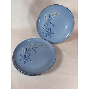 May include: A set of round ceramic plates in a light blue colour, featuring a dark blue and white wheat stalk graphic design. The plates have a smooth finish and a circular shape, with one plate standing upright and two plates stacked flat below it.