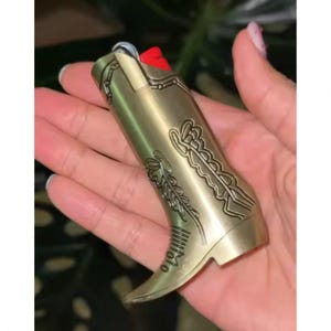 Puede incluir: Un encendedor de metal dorado con forma de bota de vaquero. La bota presenta intrincados diseños en relieve, incluyendo patrones florales y geométricos. El encendedor tiene una punta puntiaguda y un pequeño acento rojo.