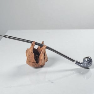 Puede incluir: Una larga pipa para fumar negra con un cuenco moteado y detalles plateados descansa sobre un soporte de madera natural. El caño de la pipa es delgado y el cuenco tiene un aspecto veteado. El soporte de madera es de color marrón claro.