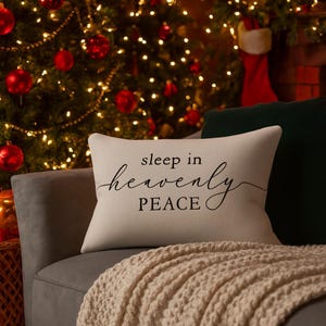 Puede incluir: Un cojín rectangular beige con las palabras "sleep in heavenly PEACE" en escritura negra. El cojín está sobre un sofá gris con una manta de punto color crema. Un árbol de Navidad con adornos rojos y luces en el fondo.