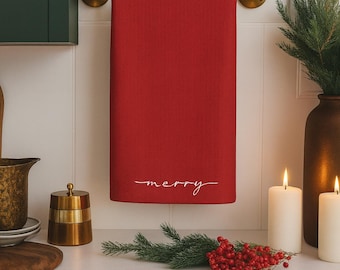 Puede incluir: Una toalla de cocina roja con la palabra «merry» en escritura blanca cuelga de una barra de toalla de latón. La escena incluye velas, vegetación y un recipiente de latón, lo que sugiere un tema festivo.