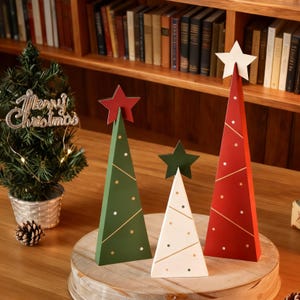 May include: Three wooden Christmas tree decorations in green, white, and red, each topped with a star. The trees have gold and white dot and line accents. A small Christmas tree with a "Merry Christmas" sign is also present.