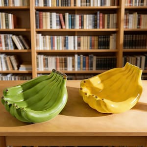 May include: Two ceramic banana-shaped bowls, one green and one yellow, sit on a wooden table. The bowls have a glossy finish and are designed to resemble a bunch of bananas. The background features a bookshelf filled with books.