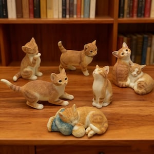 Könnte beinhalten: Eine Sammlung von orange-weiß gestreiften Katzenfiguren in verschiedenen Posen, darunter stehend, sitzend und kuschelnd. Die Figuren sind auf einer Holzoberfläche ausgestellt, mit einem Bücherregal im Hintergrund. Die Katzen haben detaillierte Merkmale und sind aus Keramik.