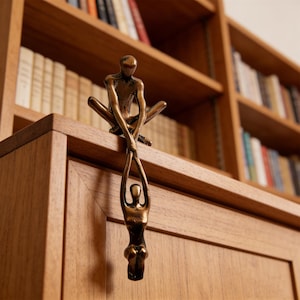 Könnte beinhalten: Bronzefarbene Skulptur zweier Figuren, eine sitzend und die andere darunter hängend. Die sitzende Figur befindet sich auf einem Holzschrank, im Hintergrund ein Bücherregal. Die Skulptur ist ein dekoratives Zierelement.