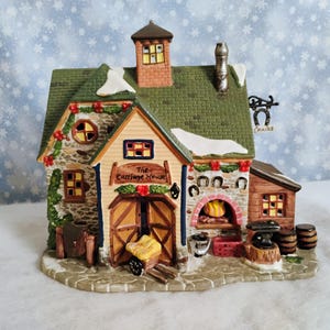 May include: A detailed ceramic miniature of "The Carriage House" with a green roof, stone walls, and wooden doors. The building features a chimney, a horseshoe sign, and decorative elements, creating a festive holiday scene. A small cart is in front of the doors.